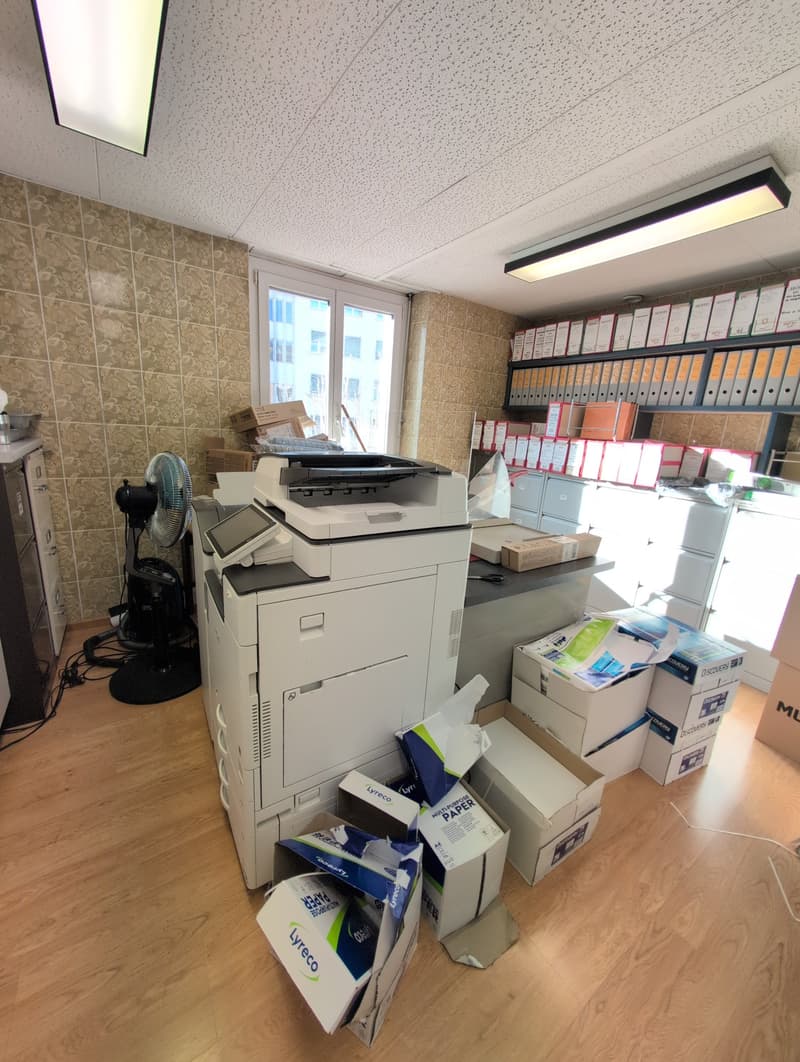 office with large printer, wooden floor, many boxes and storage boxes, large window, cabinets, fan