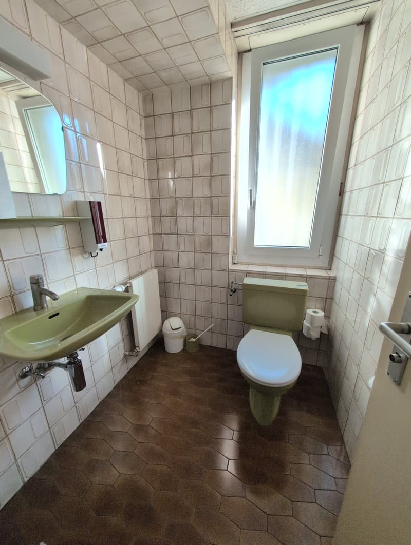 A small bathroom with tile walls, brown floor, a sink, a toilet, a mirror, a toilet paper holder, and a window.
