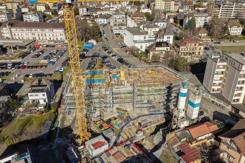 Construction site of a new building, cranes, scaffolding, multiple cars parked in surrounding areas, nearby urban landscape