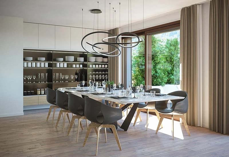 Large open-concept dining area with modern pendant lighting, wooden dining table and chairs, built-in shelving with glassware, and large windows overlooking a garden or outdoor space
