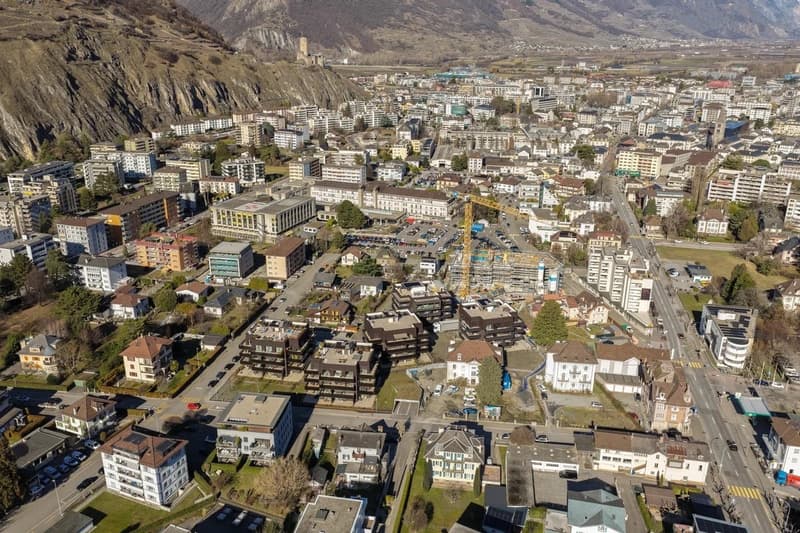 Various buildings with different architectural styles, multiple cars parked, roads, greenery, mountains in the background