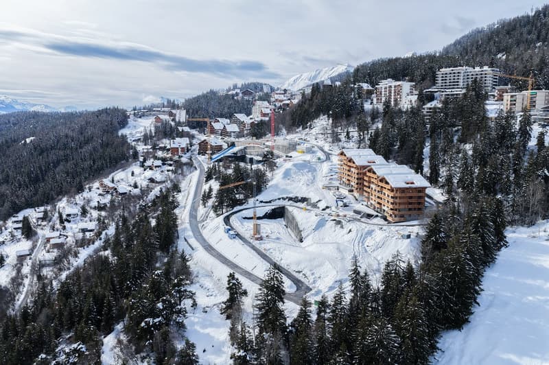 Aerial view of a ski resort with several buildings, including a large building under construction, surrounded by snow-covered trees and mountains.