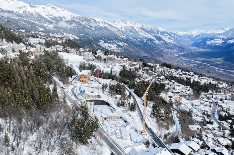 Aerial view of a mountainous region with snow-covered landscape, several buildings, and ongoing construction work.