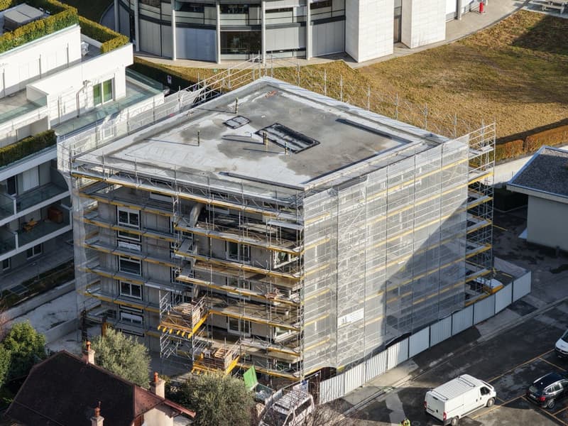Under construction multi-story building with a flat roof, windows, and scaffolding. Adjacent to other buildings and with a large open area.