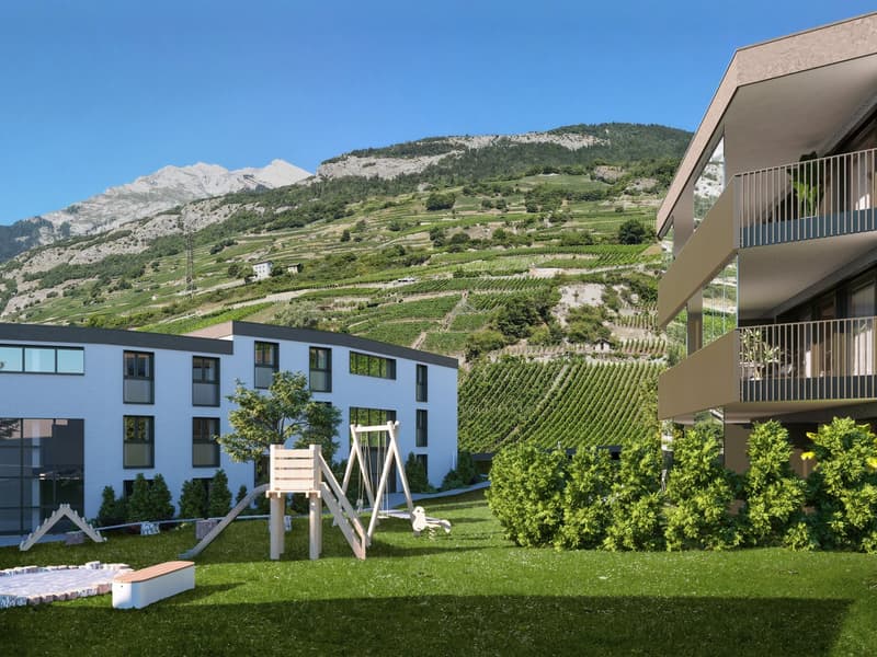 Modern apartment building with balconies, surrounded by a well-maintained garden with a playground, set against a backdrop of terraced vineyards and mountains.