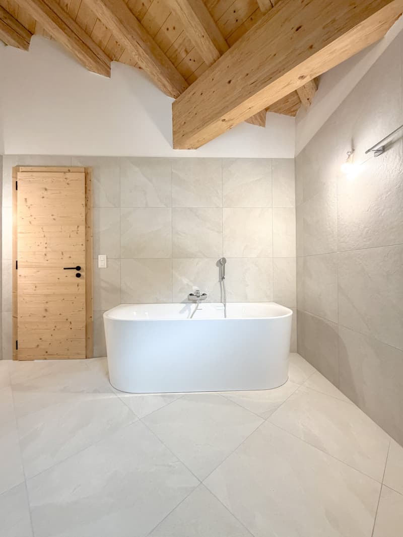 Modern bathroom with wooden ceiling, white bathtub, tile flooring, wooden door, and wall-mounted lamp