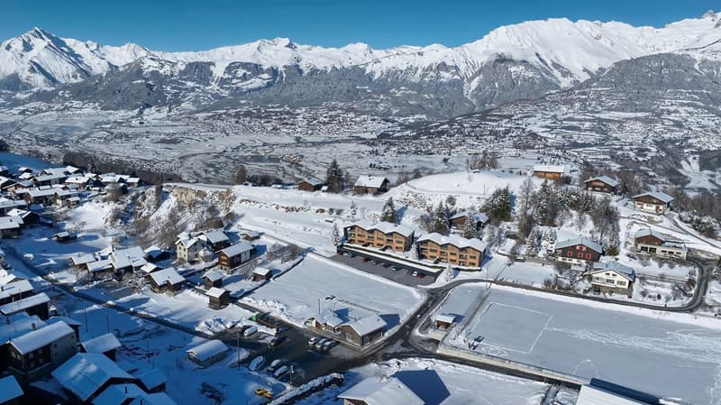 Mountainous region with several chalet-style buildings, covered in snow, roads and parking areas visible, distant snowy mountains