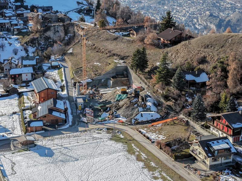 Aerial view of a hillside construction site with several buildings in progress, cranes, construction vehicles, and a large excavation area. The site is surrounded by other buildings, some with snow on the roofs, and it is set in a snowy landscape with a c