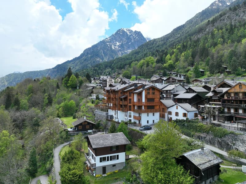 Picturesque alpine village nestled in a valley surrounded by snow-capped mountains, with traditional wooden chalets and buildings, lush green forests, and a scenic landscape.