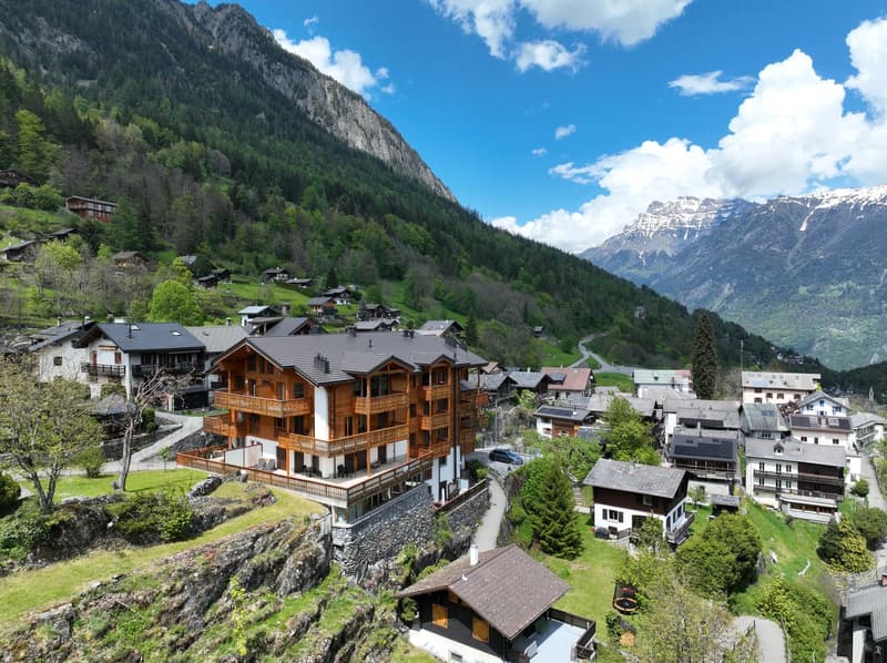 Picturesque mountain village nestled in a valley surrounded by towering snow-capped peaks and lush green forests. The village features traditional wooden chalets with balconies, a winding road, and a scenic landscape with a clear blue sky and fluffy white