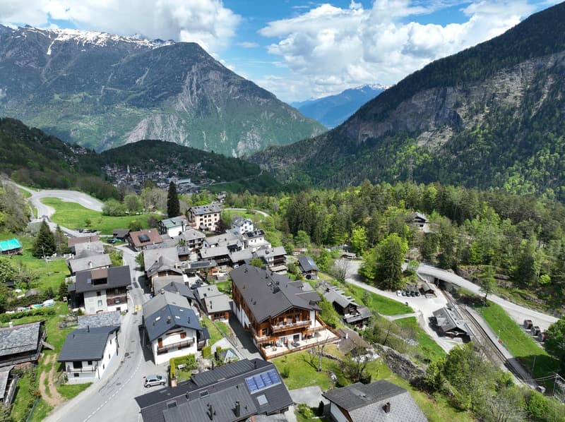 The image shows a picturesque mountain village nestled in a lush, green valley surrounded by towering snow-capped peaks. The village features a mix of traditional alpine-style buildings with balconies and terraces, as well as some modern structures. The l