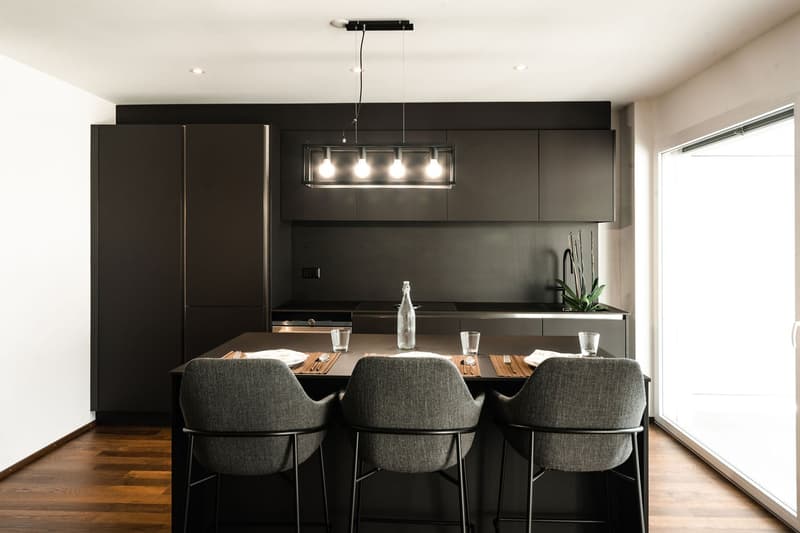 modern kitchen with black cabinets, wooden floor, large island with bar stools, pendant light, glass tableware, plant on countertop