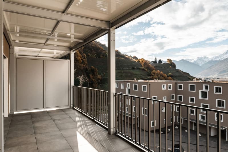 Balcony with metal railings, tiled floor, glass roof, view of mountains and buildings