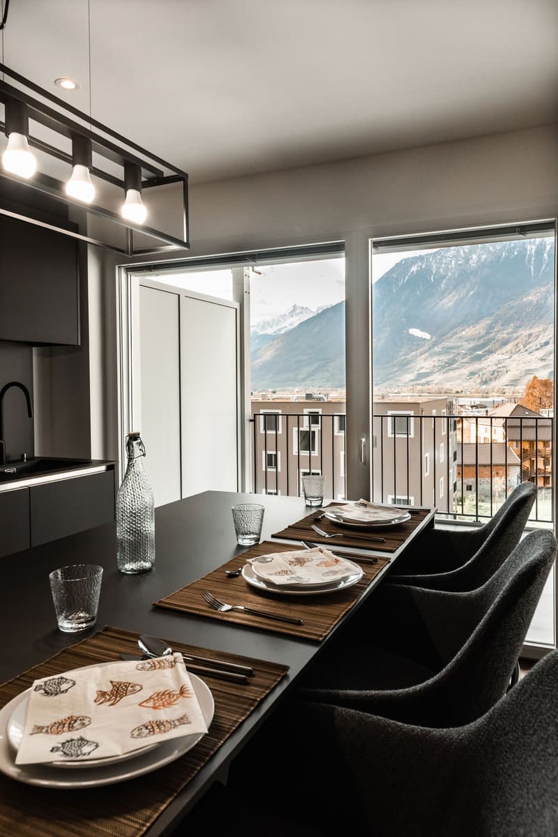 Dining table with chairs, glass bottles, plates, and cutlery. Large windows with a view of the mountains.