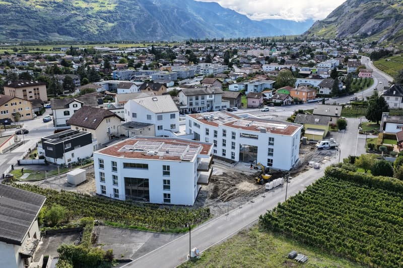 Aerial view of a modern apartment building under construction, surrounded by other buildings and a scenic mountain backdrop.