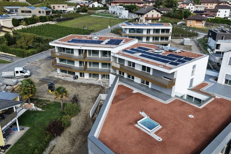 Aerial view of a modern apartment complex with solar panels on the roofs, a swimming pool on the terrace, and a construction site in front.