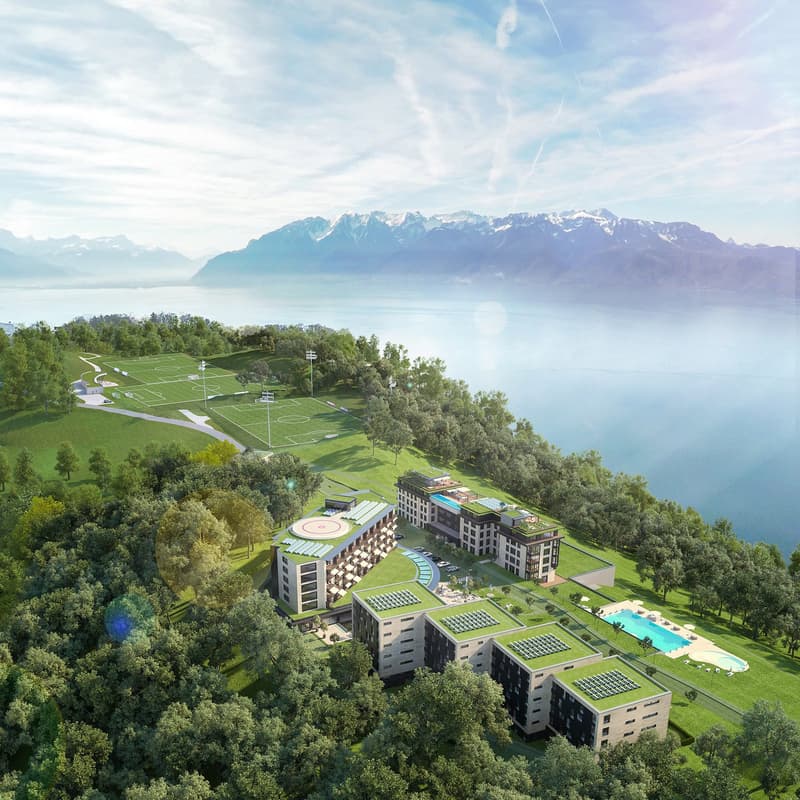 Aerial view of a resort with multiple buildings, swimming pool, and a view of the mountains and lake