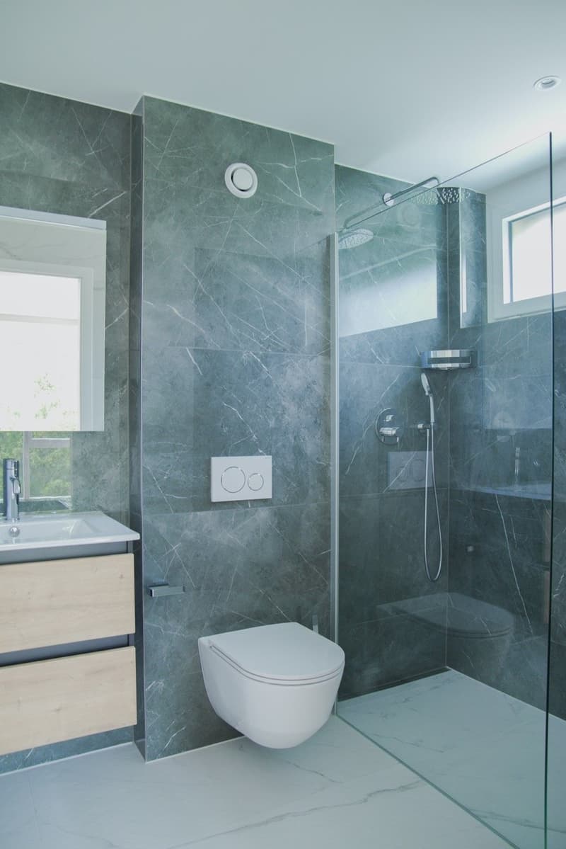 modern bathroom with gray marble walls, white toilet, glass shower enclosure, white sink, wooden cabinet, silver fixtures
