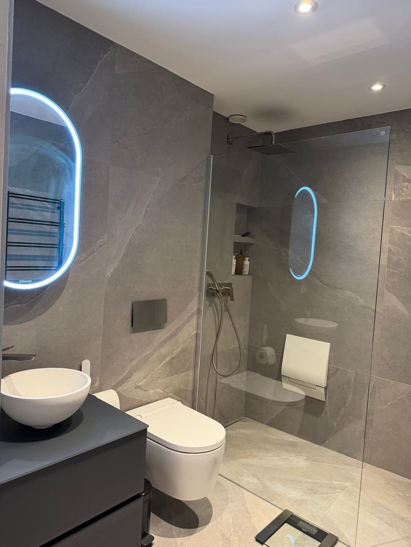 modern bathroom with gray tiles, white toilet, white sink, glass shower enclosure, illuminated mirror, built-in shelves