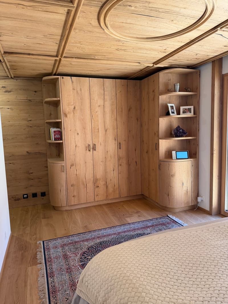 Bedroom with wooden walls, wooden floor, large wooden wardrobe, bed, rug, shelves with books and decorative items