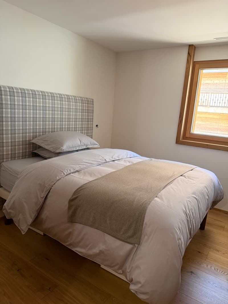 Bedroom with a bed, plaid headboard, two pillows, white bedding, wooden floor, white walls, window with wooden frame