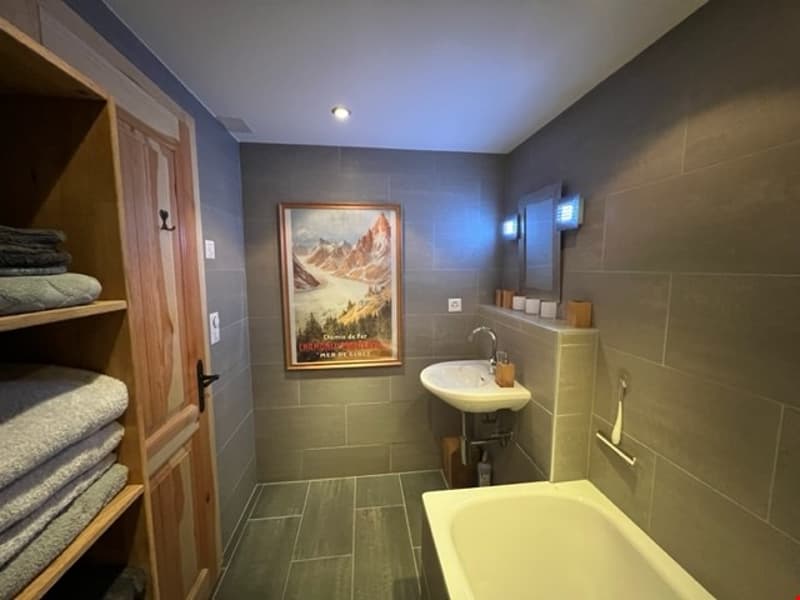 The image shows a spacious bathroom with gray tile walls and floors. There is a large bathtub, a sink with a vanity, and shelving units for storage. The room also features a framed artwork on the wall, adding a decorative touch to the space.