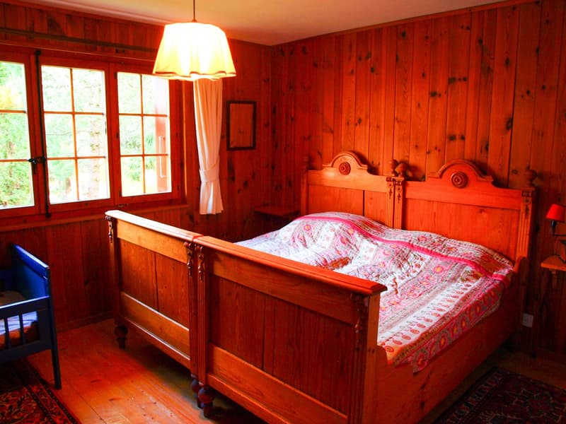 Rustic wooden bedroom with a large wooden bed frame, patterned bedding, and a hanging lamp. The room has wooden paneled walls and large windows overlooking the outdoors.