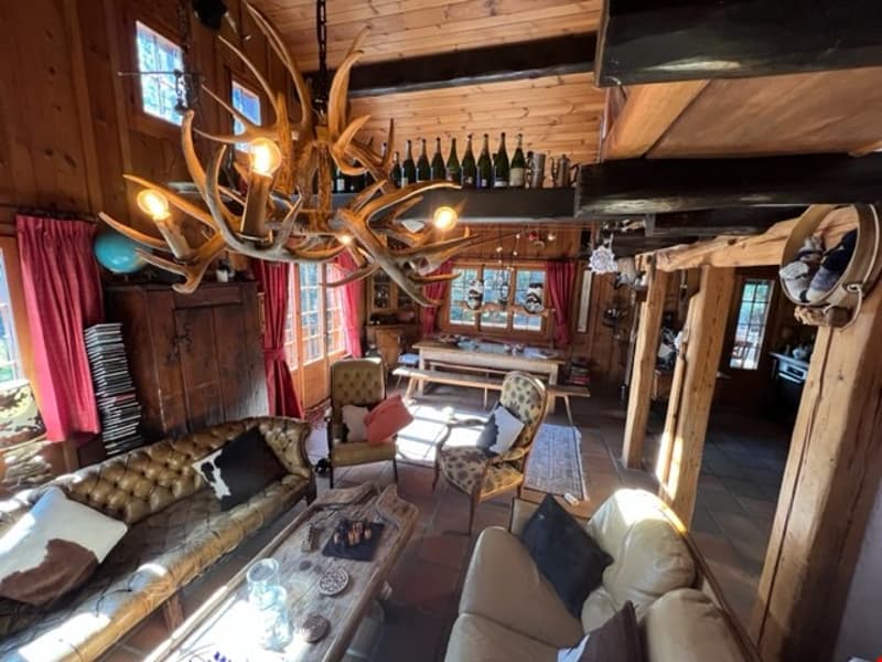 Rustic log cabin interior with wooden walls and ceiling, antler chandelier, leather and upholstered furniture, fireplace, and wine bottles displayed on shelves.