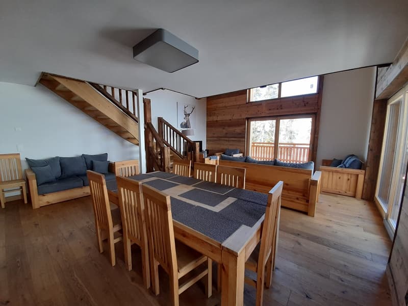 Wooden dining table and chairs, cozy living room with a couch and a staircase, wooden flooring, glass windows, wooden decorative pieces