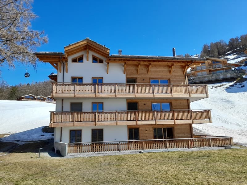 wooden apartment building with three balconies, wooden fencing, white exterior, red roof, snowy mountainside in background