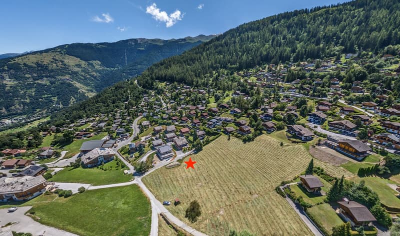 Aerial view of a mountainous village with wooden chalets, an empty plot of land with a red star, surrounded by trees and mountains