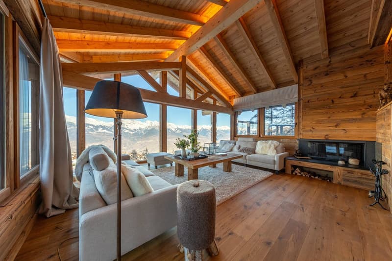Open plan living room with wooden floors, wooden ceiling beams, fireplace, panoramic windows with mountain view, wooden coffee table, white sofas, lamps, rug, ottoman, floor-standing plant