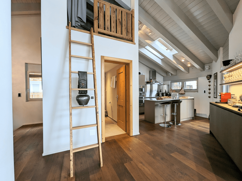 Open-plan kitchen, dining, and living area with wooden floor and high ceilings, small ladder leading to an attic, kitchen island with bar stools, and a skylight.