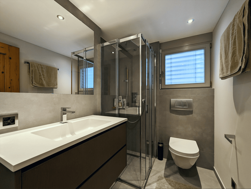 gray tiled walls, glass walk-in shower, white sink, white toilet, wooden floors, mirror, wooden door