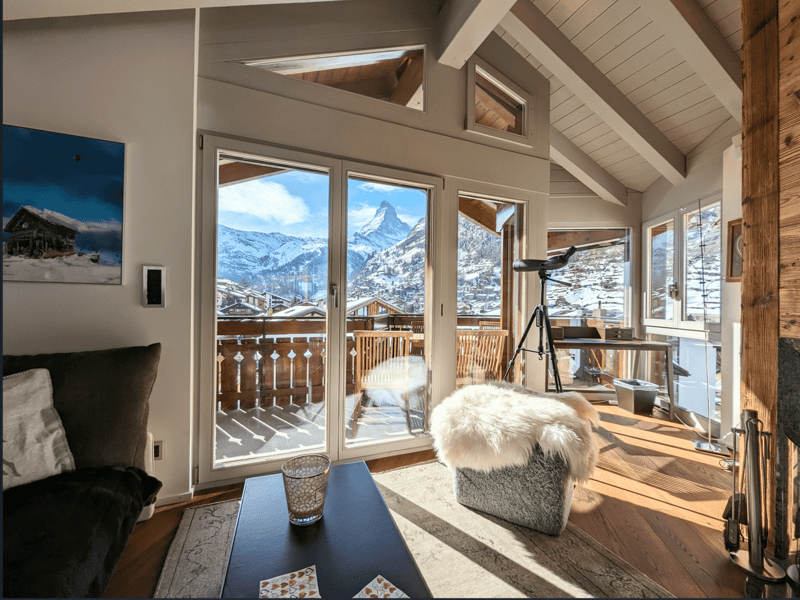 Glass doors leading to balcony, wooden flooring, mountains visible in the background, fireplace, sofa, wooden table, table top with wine bottle, telescope