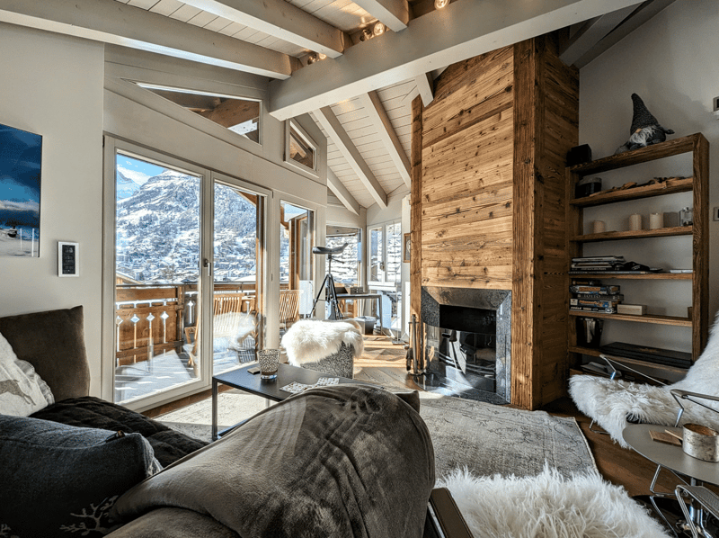 large glass wall, modern fireplace, wooden beams, balcony with snowy mountain view, fur chair, wooden table, rug, sofa