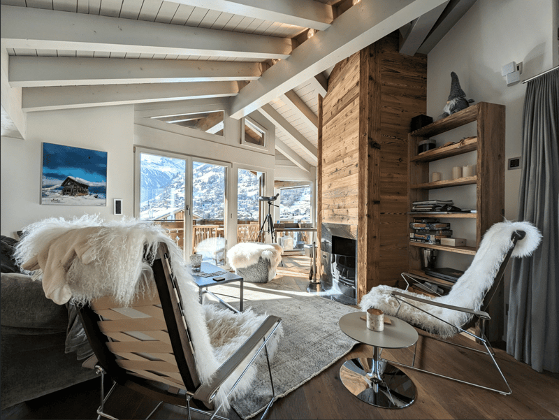 Wooden beams, large windows with mountains in the distance, wooden floor, wooden cabinet, fireplace, couches, tables, chairs with fur seat and backrest