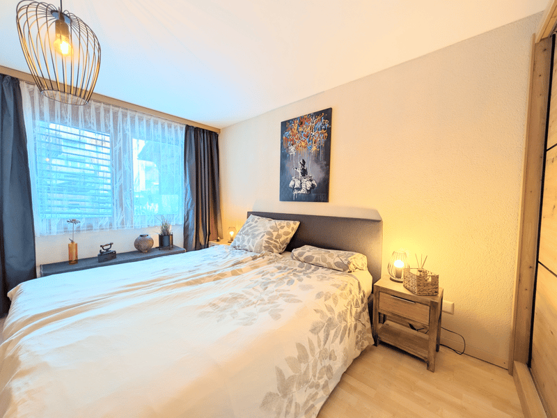 Bedroom with a double bed, nightstands, a window with curtains, a painting on the wall, and a shelf with decorative items.