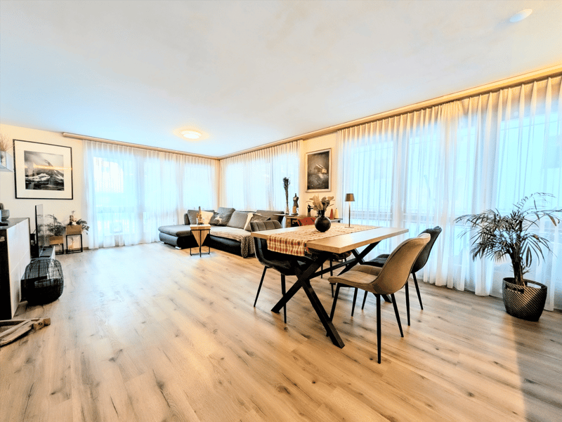 Open floor plan, hardwood floors, large windows, dining table, couch, side table, potted plants, picture frames, ceiling lamp