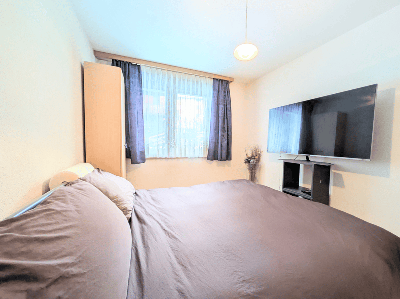 Bedroom with a double bed, a cabinet, a TV, a window with curtains, and a vase with flowers on a shelf.