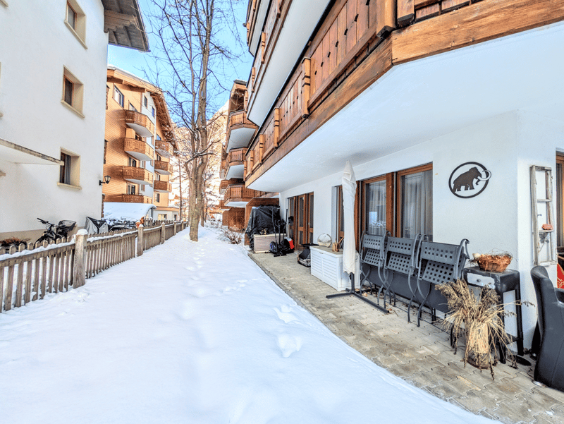Snowy alley between two buildings, wooden fence, bicycles, outdoor furniture, potted plants, wooden balconies, snow on the ground
