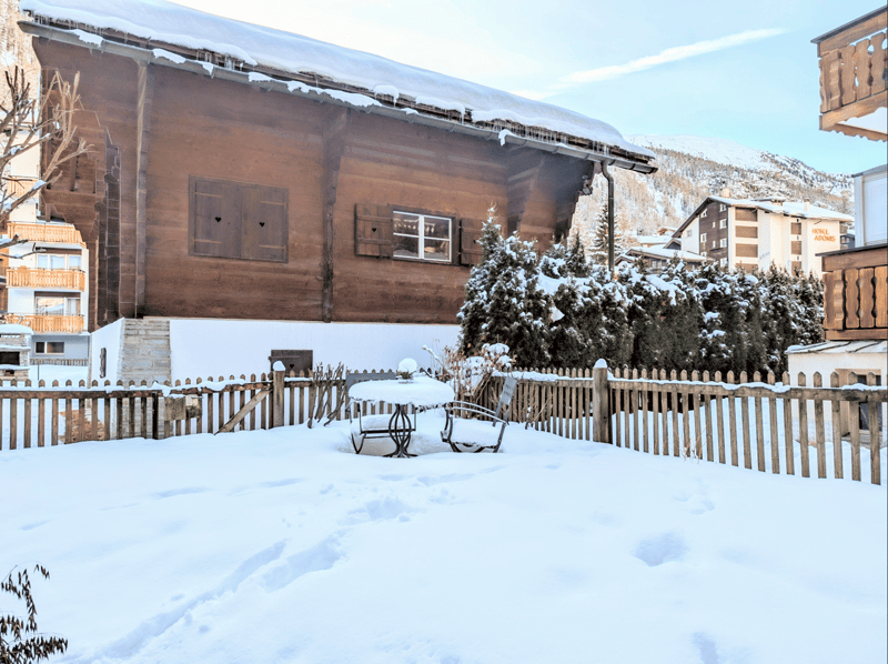 wooden house, snow on the roof, wooden fence, table and chairs in the snow, pine trees, mountains in the background