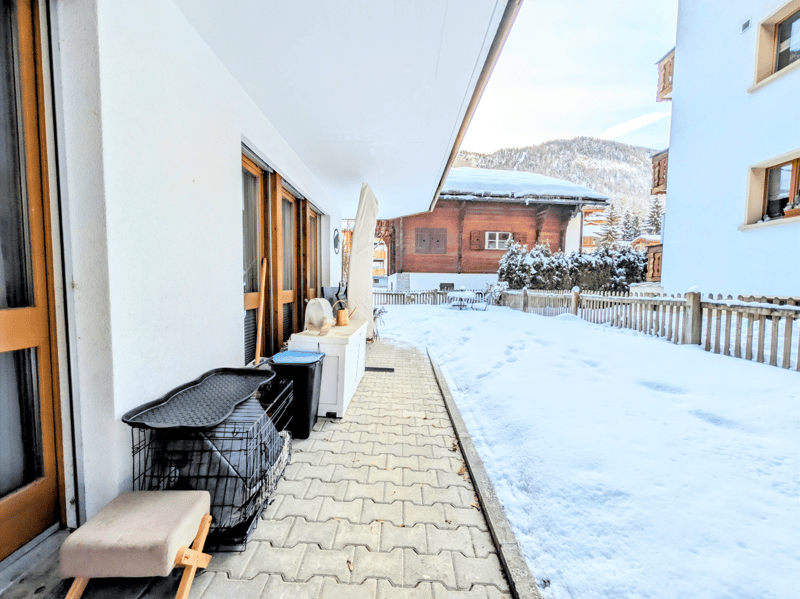 A covered walkway with a brick floor leading to a balcony with a wooden door, a bench, and a storage box. The walkway is surrounded by snow, and there is a wooden fence on the right side. In the distance, there is a house with a balcony and a snow-covered