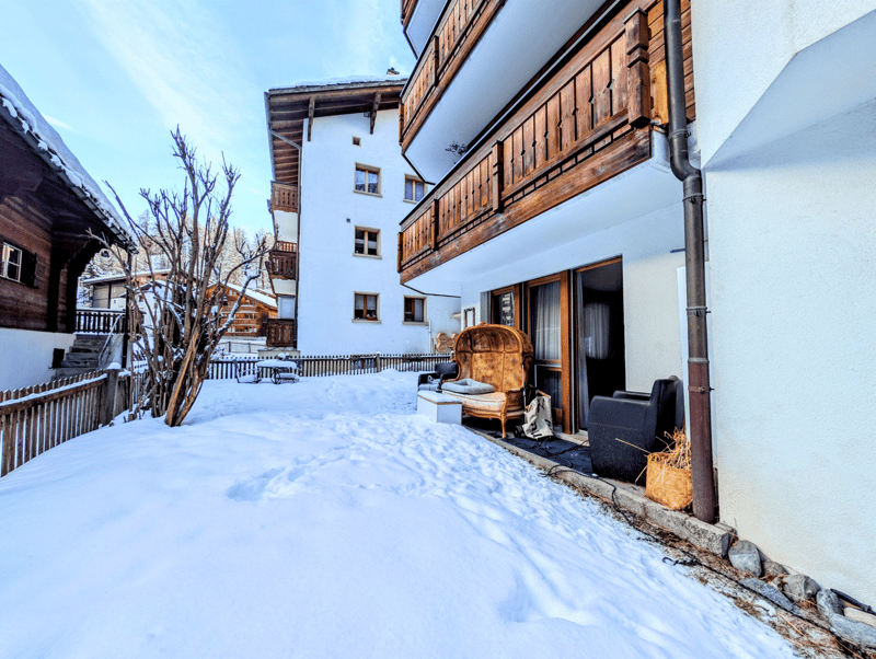 white building with wooden balconies, snow on the ground, wooden fence, tree, outdoor furniture
