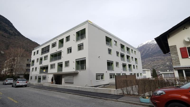 Multi-story apartment building with gray exterior, balconies, and windows. The building is situated in a mountainous area with other buildings and vehicles visible in the surroundings.