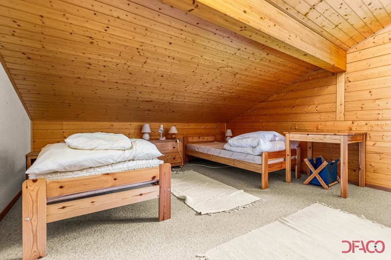 Small attic room with wooden walls and ceiling, two wooden beds, nightstands, carpet, and a small table with a blue basket. Nightstands feature lamps.