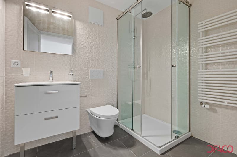 Modern bathroom with white vanity, toilet, and glass-enclosed shower. The walls are tiled with a light-colored mosaic pattern. The bathroom also features a mirror and lighting fixtures.