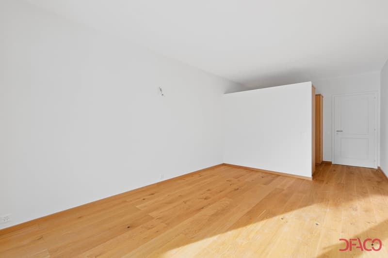 Empty room with white walls and hardwood flooring. The room appears to be a living room or bedroom with a door visible in the corner.