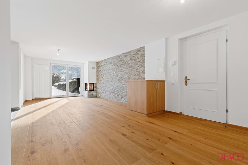 Spacious living room with hardwood flooring, a stone accent wall with a fireplace, large windows overlooking a snowy outdoor scene, and a wooden cabinet or storage unit.