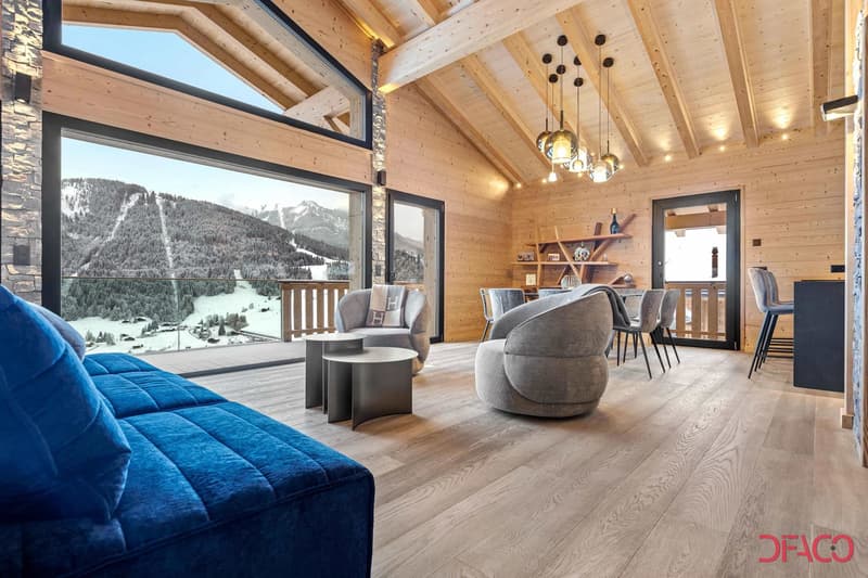 Spacious open-concept living area with high vaulted ceilings, large windows offering a scenic mountain view, wooden walls and floors, comfortable gray sofas and chairs, a dining table, and modern pendant lighting fixtures.